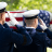 Three uniformed military personnel salute a flag-draped coffin during a solemn outdoor funeral.